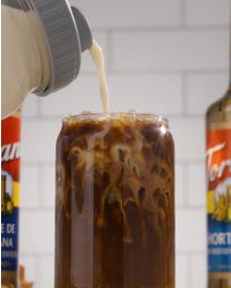 Iced coffee topped with cream on a table next to syrup bottles.