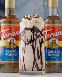 Banana split milkshake in a glass topped with bananas, nuts, chocolate sauce.