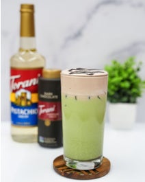 Dubai Chocolate Latte in a glass in front of syrup and sauce bottle.