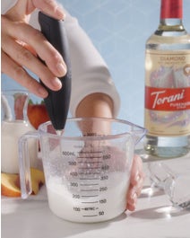 Person frothing mixture in measuring cup next to a bottle of diamond syrup.