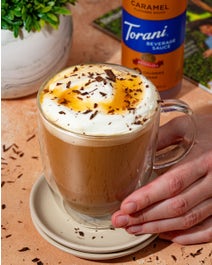 Sugar Free Caramel Latte in a glass mug being held on a table in front of sauce bottle.