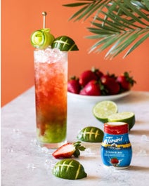 Touchdown Strawberry Cooler in a glass on a table with fruit and drink enhancer on a table.